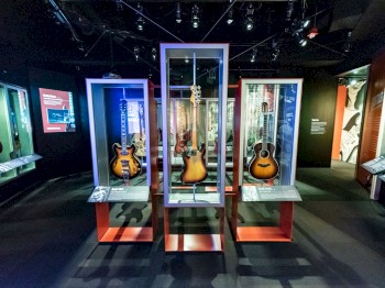 The image shows a museum exhibit featuring various guitars displayed in glass cases under spotlights, with informative panels on the walls.