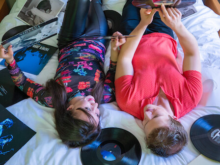 Two kids lying on a bed, vinyl records around them, one with a tablet; relaxed, casual movie-night vibe.