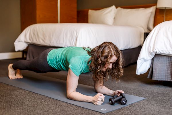 A person is doing a plank exercise on a yoga mat in a room with beds. They are wearing a green shirt and black pants, holding a small weight.