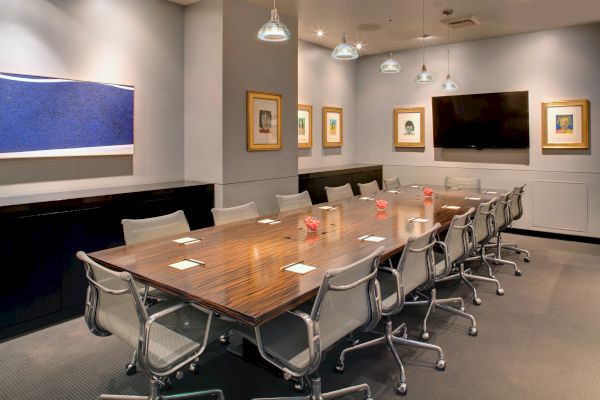 This image shows a modern conference room with a long wooden table, multiple chairs, framed art on the walls, and a large TV.