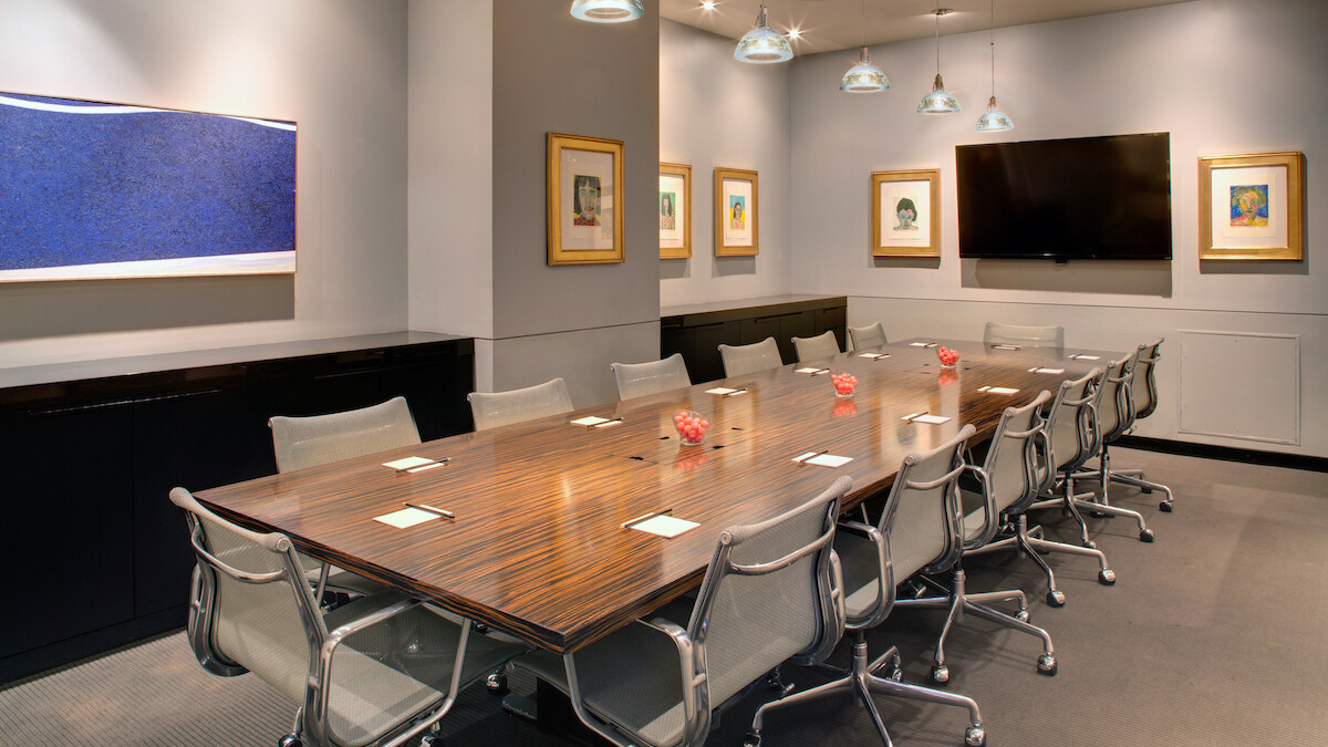 A modern conference room with a long table, chairs, artwork on the walls, and a large TV screen, illuminated by several hanging lights.