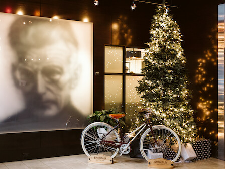 A dimly lit display with a large blurred portrait, festive Christmas tree, warm string lights, and two vintage bikes beside wrapped gifts.