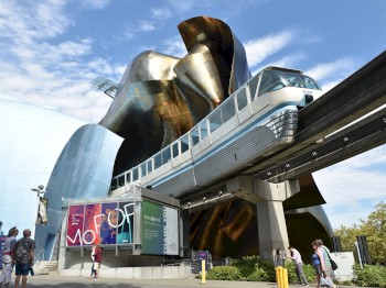 A futuristic structure with a monorail passing by, people walking, and colorful signage on a modern building under a partially cloudy sky.