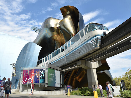 A futuristic structure with a monorail passing by, people walking, and colorful signage on a modern building under a partially cloudy sky.