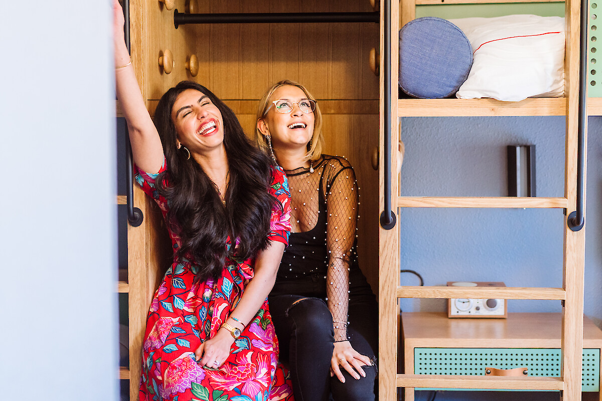 Two women sit smiling in a doorway of a cozy, bright room with shelves and cushions overhead, enjoying a playful, sunny moment.