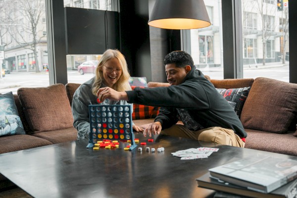 Two people are playing a board game on a coffee table in a cozy, modern living room with large windows and a lamp overhead.