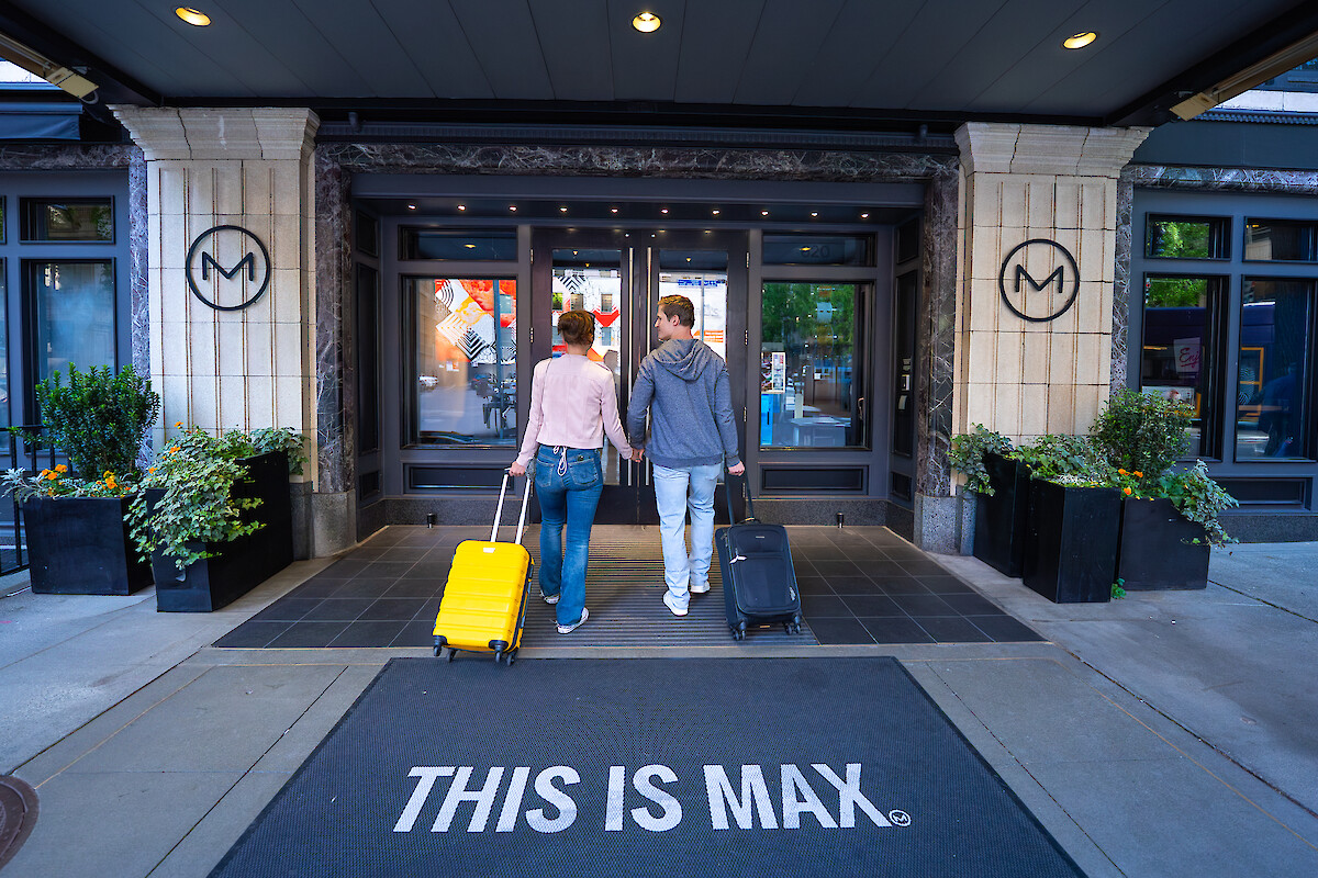 A couple enters a building, each with a suitcase, under a sign reading 