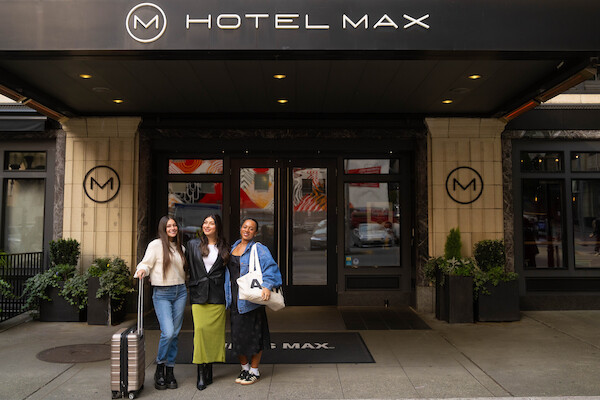 Three people with luggage stand in front of Hotel Max, posing for a photo near the entrance.