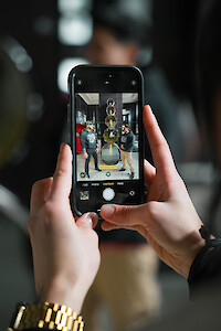 A person holding a phone, taking a photo of someone in front of a tower of circular objects, with people in the background.