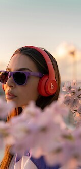 A person wearing red headphones and purple sunglasses, surrounded by pink flowers at sunset.