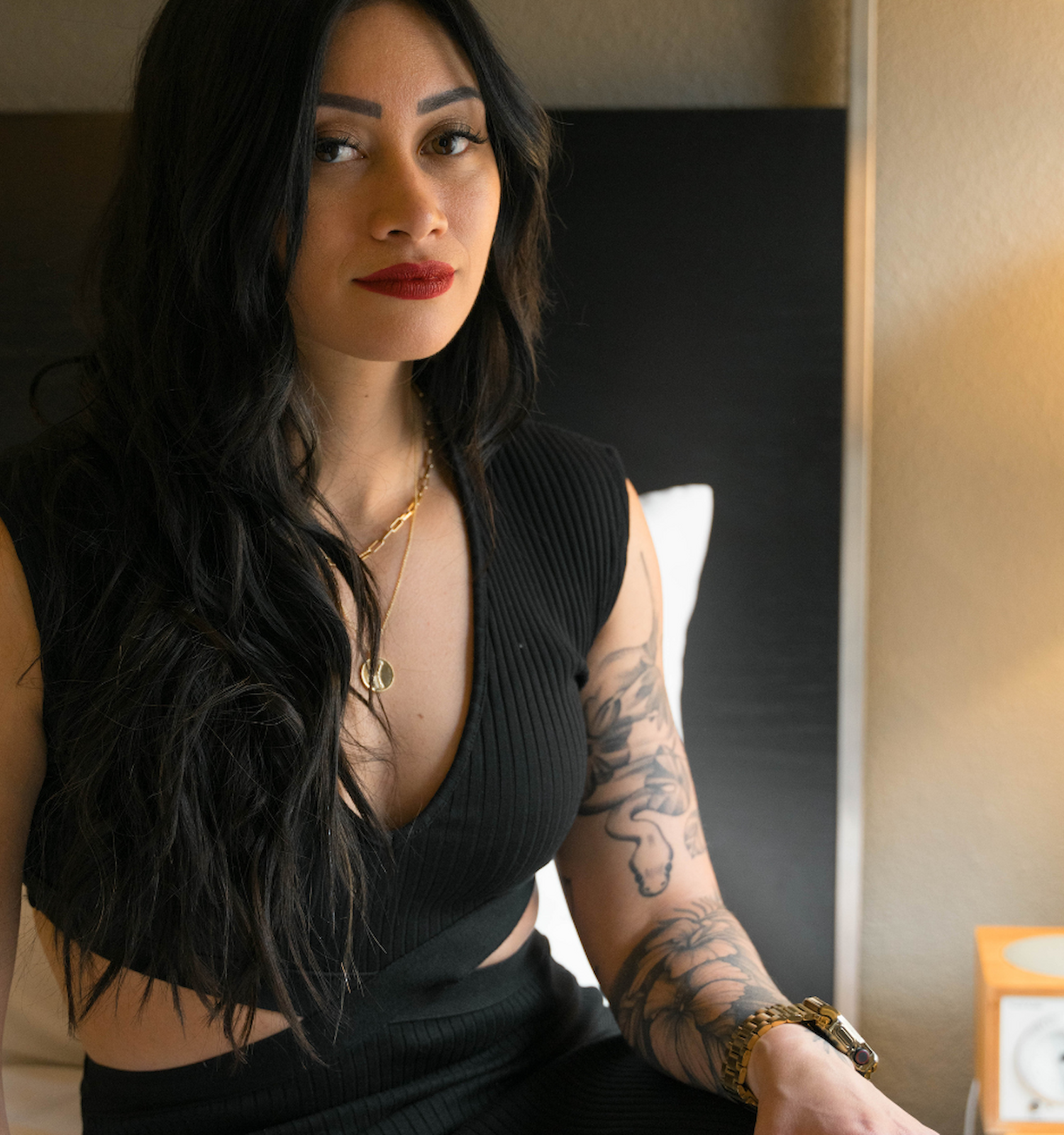 A person with long hair and tattoos sits on a bed, wearing a black top. A bedside table with a lamp is visible.