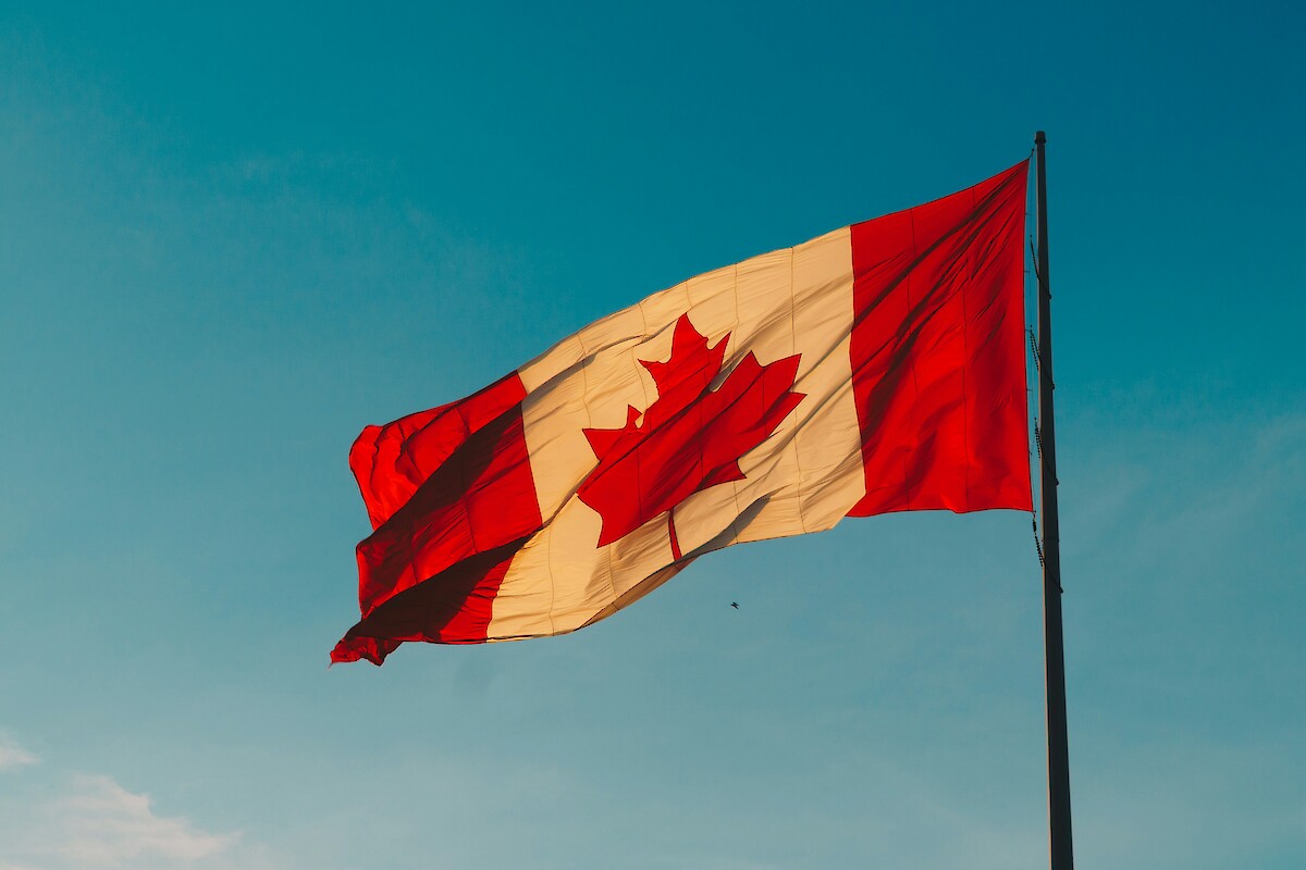 The image shows a Canadian flag with a red maple leaf centered, waving against a clear blue sky.