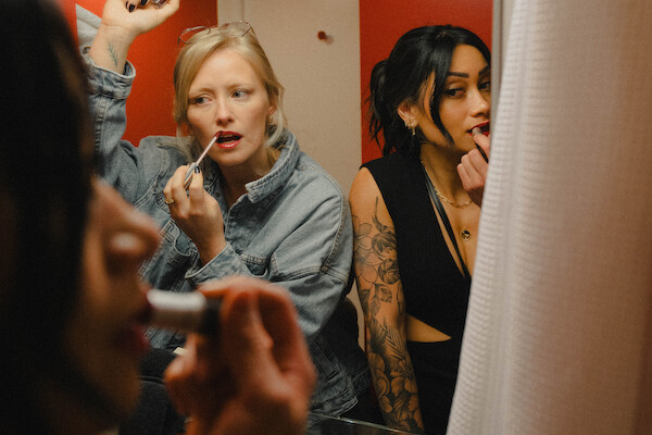 Two people are applying makeup in front of a mirror in a small, brightly lit room with red walls. One holds a brush, the other uses lipstick.