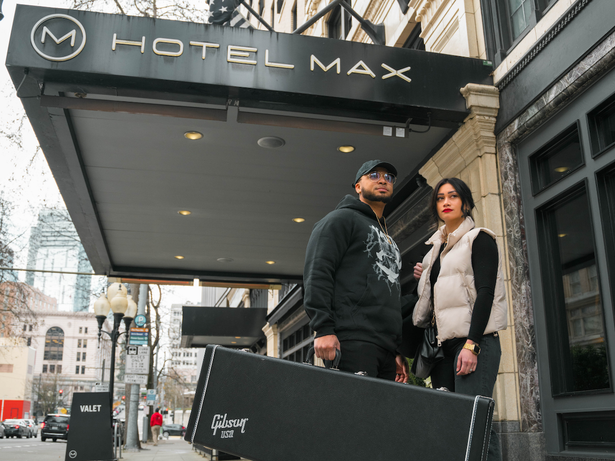 Two people stand in front of Hotel Max, one holding a Gibson guitar case, under a large canopy with the hotel sign above them.