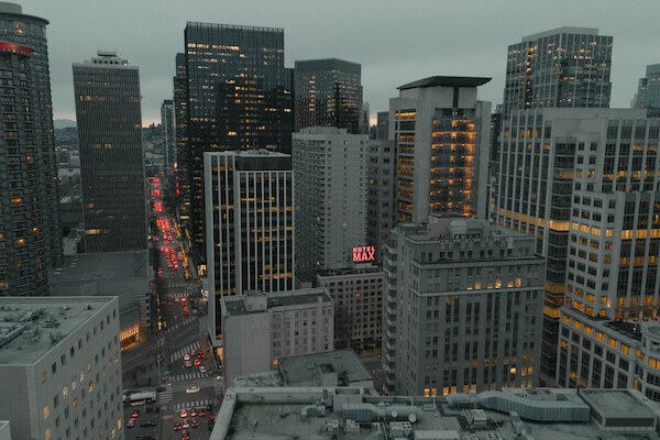 An overcast view of a cityscape with high-rise buildings and bustling streets lit up with traffic and building lights at dusk.