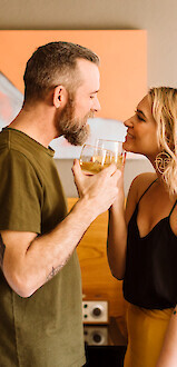 A couple stands in a room, smiling at each other while holding drinks.