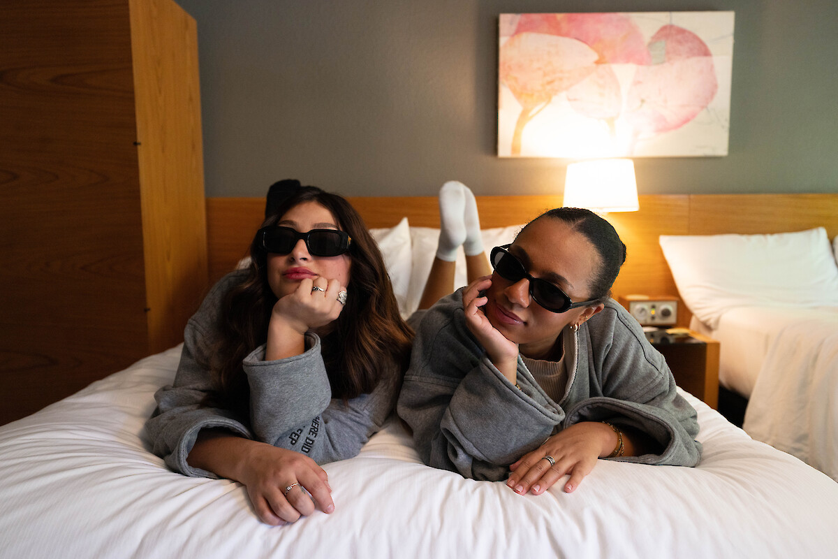 Two people wearing sunglasses and gray robes lie on a bed in a cozy room, smiling and posing casually for the camera.