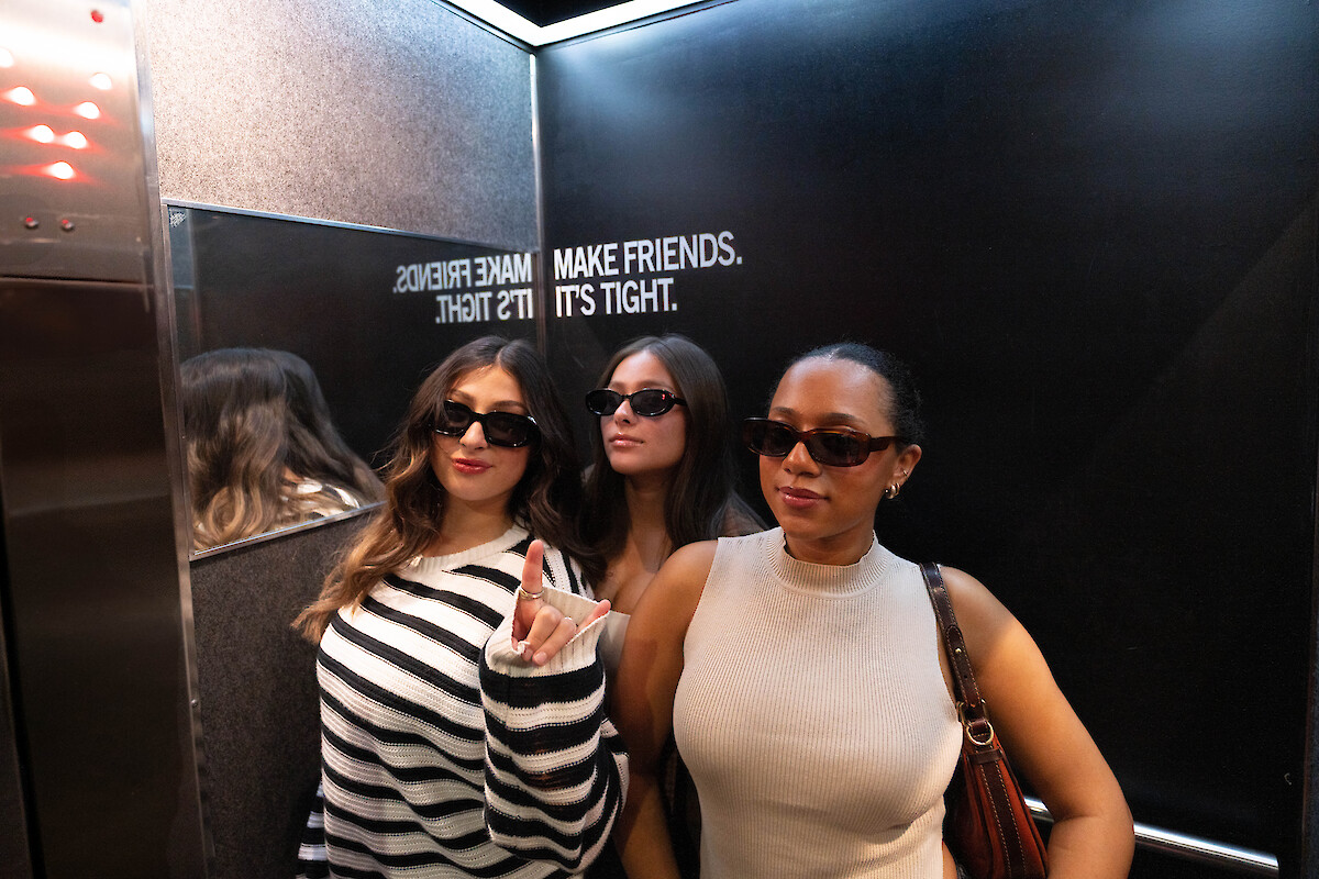 Three people wearing sunglasses pose in an elevator with a reflective wall. The text on the wall reads: 