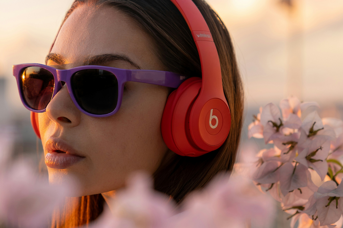 A person wearing purple sunglasses and red Beats headphones, surrounded by pink flowers, is bathed in soft sunset lighting.