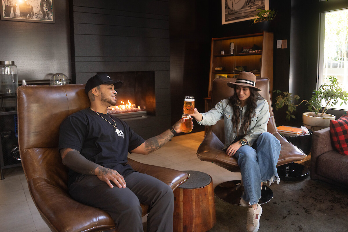 Two people are sitting in chairs near a fireplace, toasting with drinks in a cozy room.