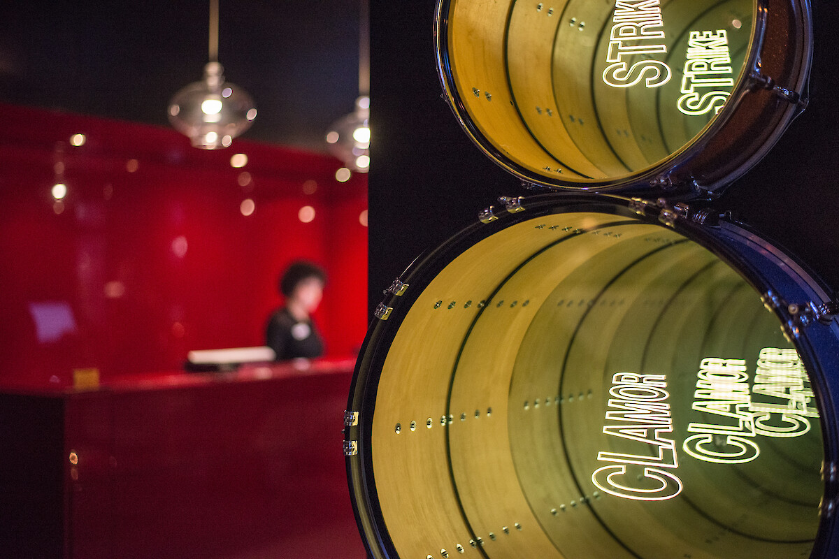 A blurred reception area with a person, red walls, hanging lights, and focus on two drums displaying the words 