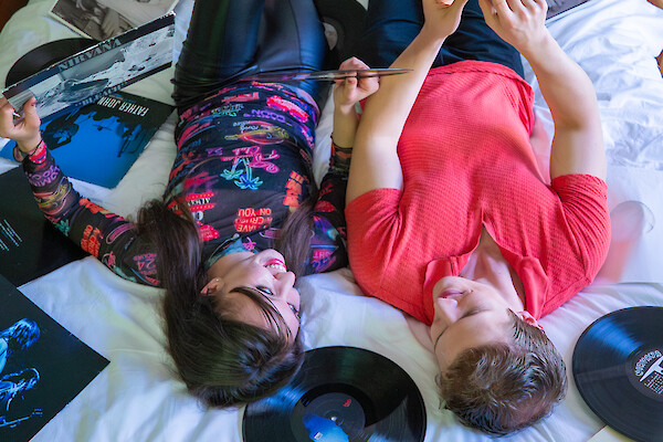 Two people lie on a bed, vinyl records around them, one using a tablet while the other watches; casual, retro vibe.