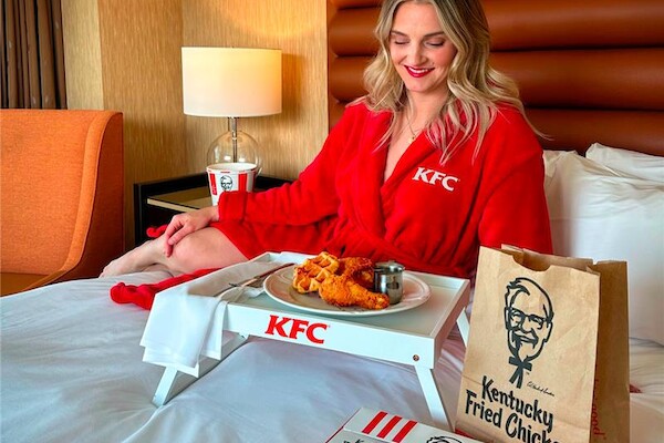 A person in a red KFC robe is enjoying fried chicken and waffles on a bed with branded KFC items around.
