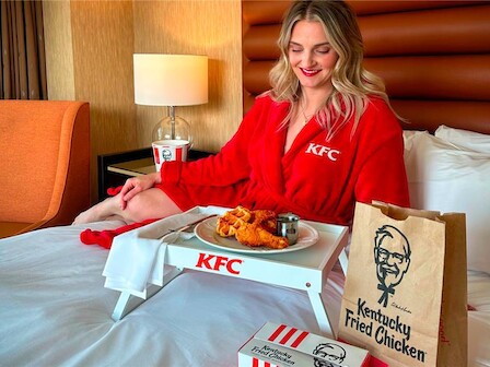 A person in a KFC robe sits on a bed with a tray of fried chicken, waffles, and a drink, beside KFC packaging, in a cozy setting.