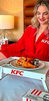 A person in a red KFC robe sits on a bed with a tray of Kentucky Fried Chicken, a bucket, and a bag of KFC food nearby.