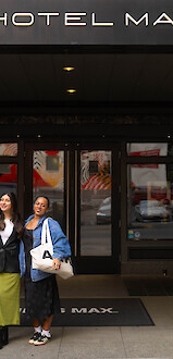 Three people stand in front of Hotel Max entrance, one with luggage, smiling and posing for a photo.