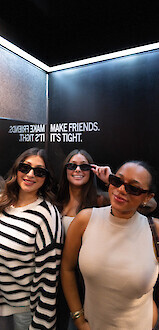 Three people wearing sunglasses, posing and smiling in a room with mirrored walls displaying text: 