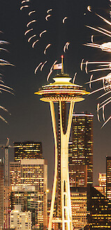 The image shows fireworks over the Seattle skyline at night, with the Space Needle prominently illuminated among tall buildings.