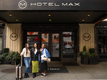 Three people with luggage stand in front of the entrance to Hotel Max, featuring a black awning and a welcoming atmosphere.