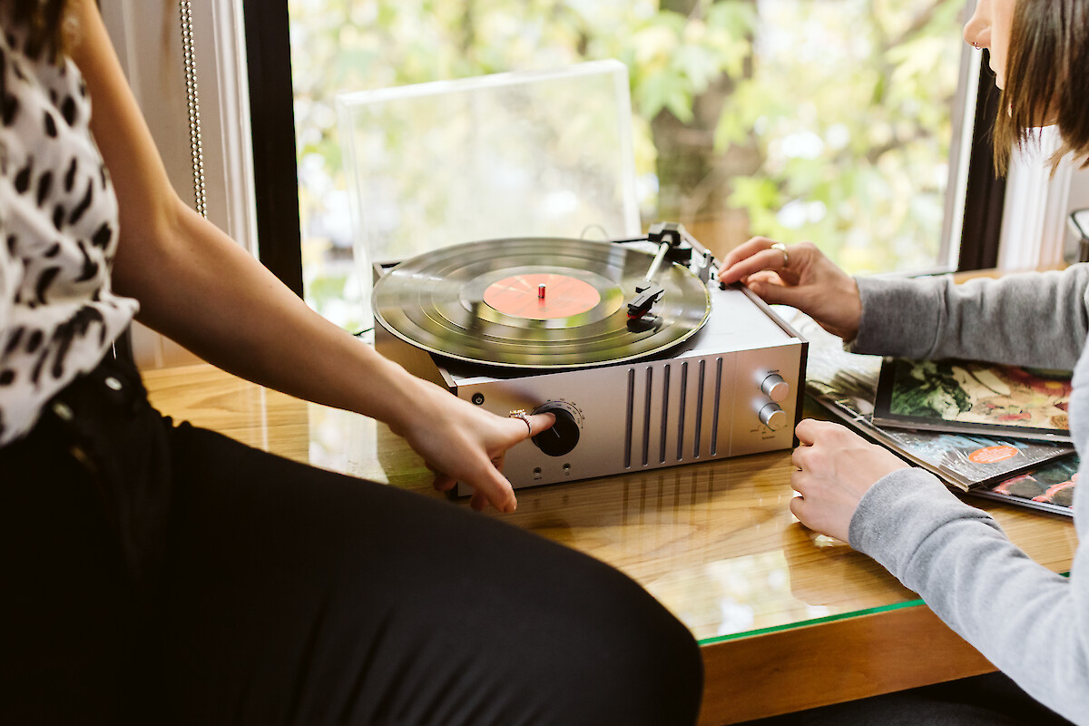 Two people are setting up a vinyl record player on a table, with records nearby and greenery visible outside a window.