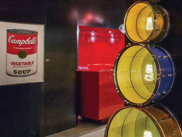 The image features a Campbell's Vegetable Soup sign and a vertical stack of colorful drums in a museum-like setting, next to a red abstract structure.