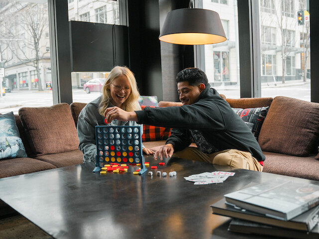 Two people are sitting on a sofa playing Connect Four in a modern living room with large windows, under a floor lamp. Books are on the table.