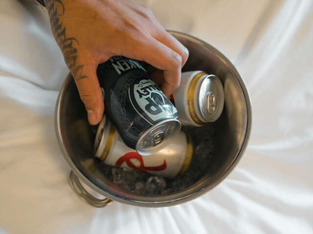 A hand with a tattoo and a wristwatch is holding cans of beer in an ice bucket on a white surface, appearing to be on a bed.