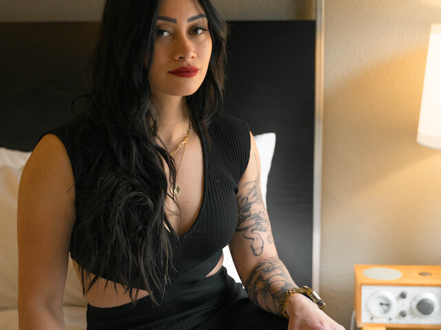 A woman with long dark hair, wearing a black dress, is sitting on a bed in a room with a bedside table and a lamp on.