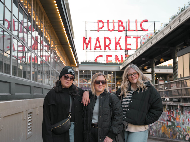 Three people are posing in front of the iconic Public Market Center sign, likely at Pike Place Market. They are dressed in casual winter clothing.