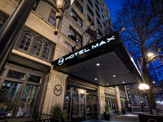 The image shows the entrance of Hotel Max, featuring a contemporary facade with signage and a well-lit walkway, set against a twilight cityscape.