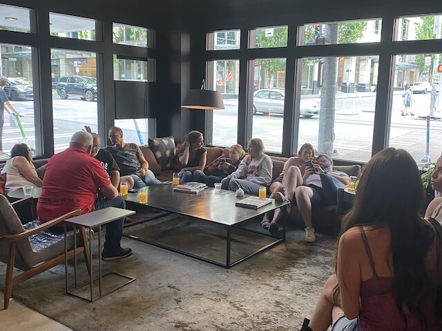 A group of people is gathered in a modern lounge with large windows, seated on couches and chairs around a coffee table.