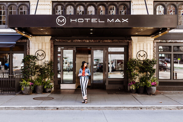 A person stands in front of Hotel Max's entrance, with potted plants on either side and decorative windows above the sign.