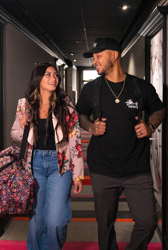 Two people walking in a hallway with bags, smiling and looking at each other. The hallway features dark walls and decorative elements.