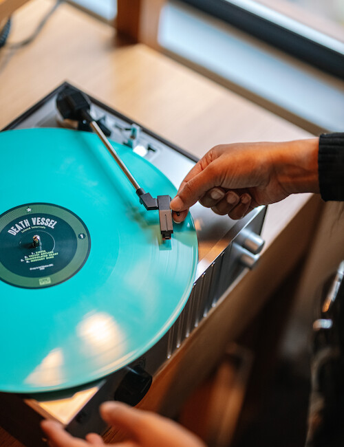 A person places a needle on a spinning turquoise vinyl record on a turntable, with the window light illuminating the scene.