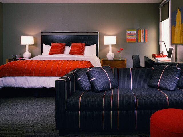 A stylish bedroom with a double bed, red linens, two lamps, a black-striped sofa, and a desk with vibrant wall art accents the decor.
