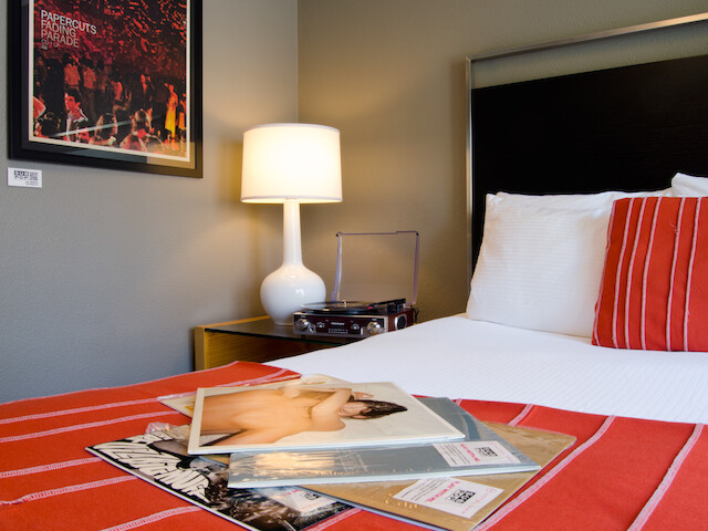 A cozy bedroom features a bed with red-striped bedding, a turntable, records, a table lamp, and framed art on the wall.