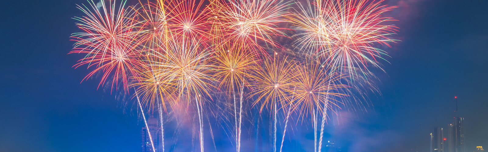 Vibrant fireworks light up the night sky over a river, with boats and a bridge in the foreground, creating a festive atmosphere.