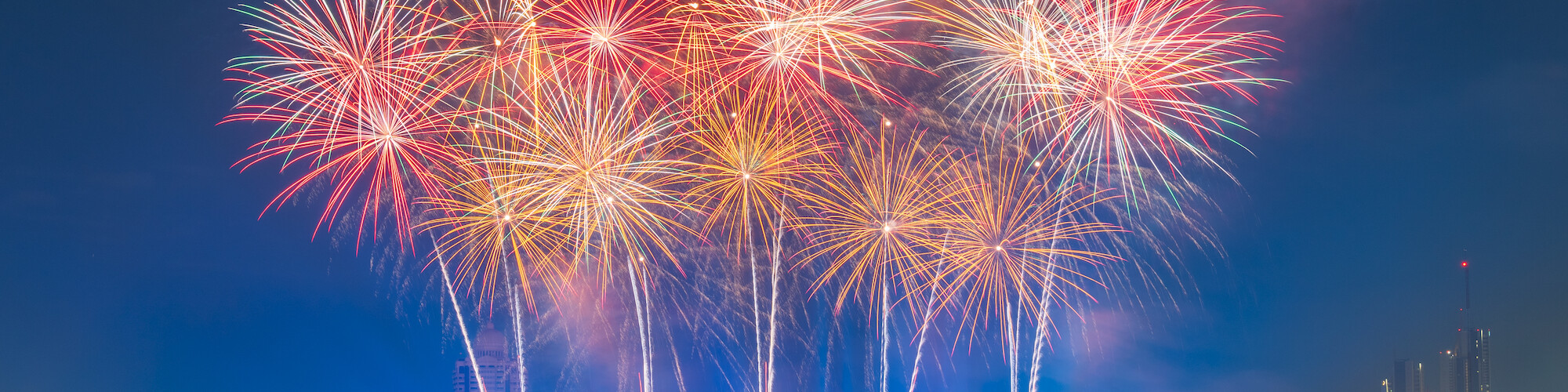 Vibrant fireworks light up the night sky over a river, with boats and a bridge in the foreground, creating a festive atmosphere.