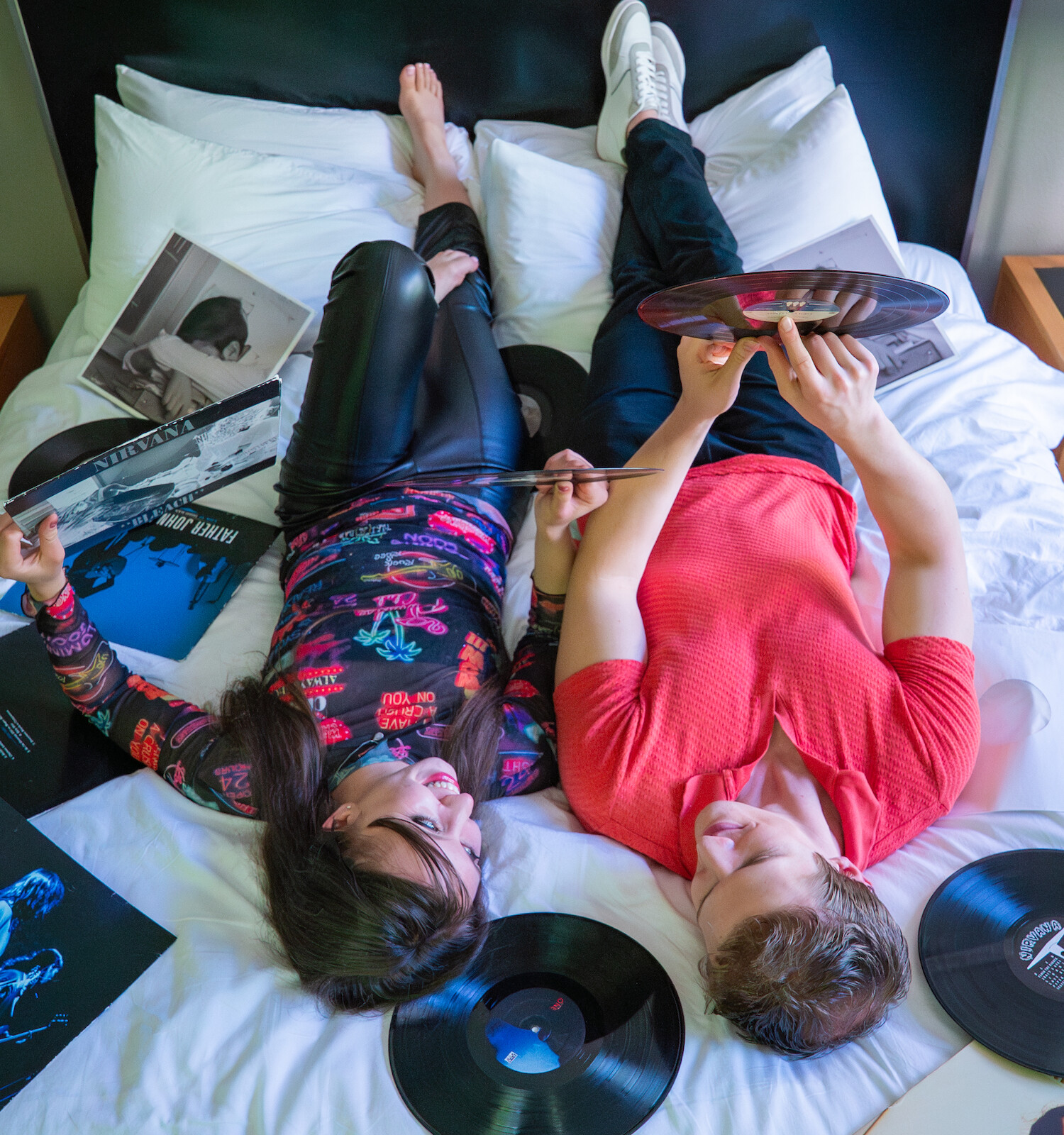 Two young people are relaxing on a bed surrounded by vinyl records and music albums, enjoying a shared moment and good music.