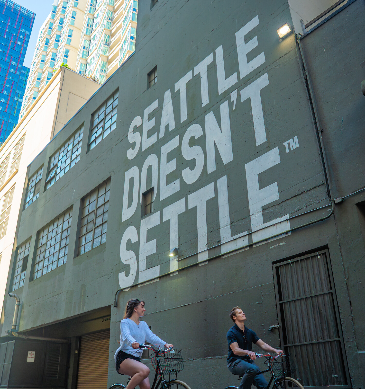 A woman and a man ride bikes in front of a wall with a big mural that reads 